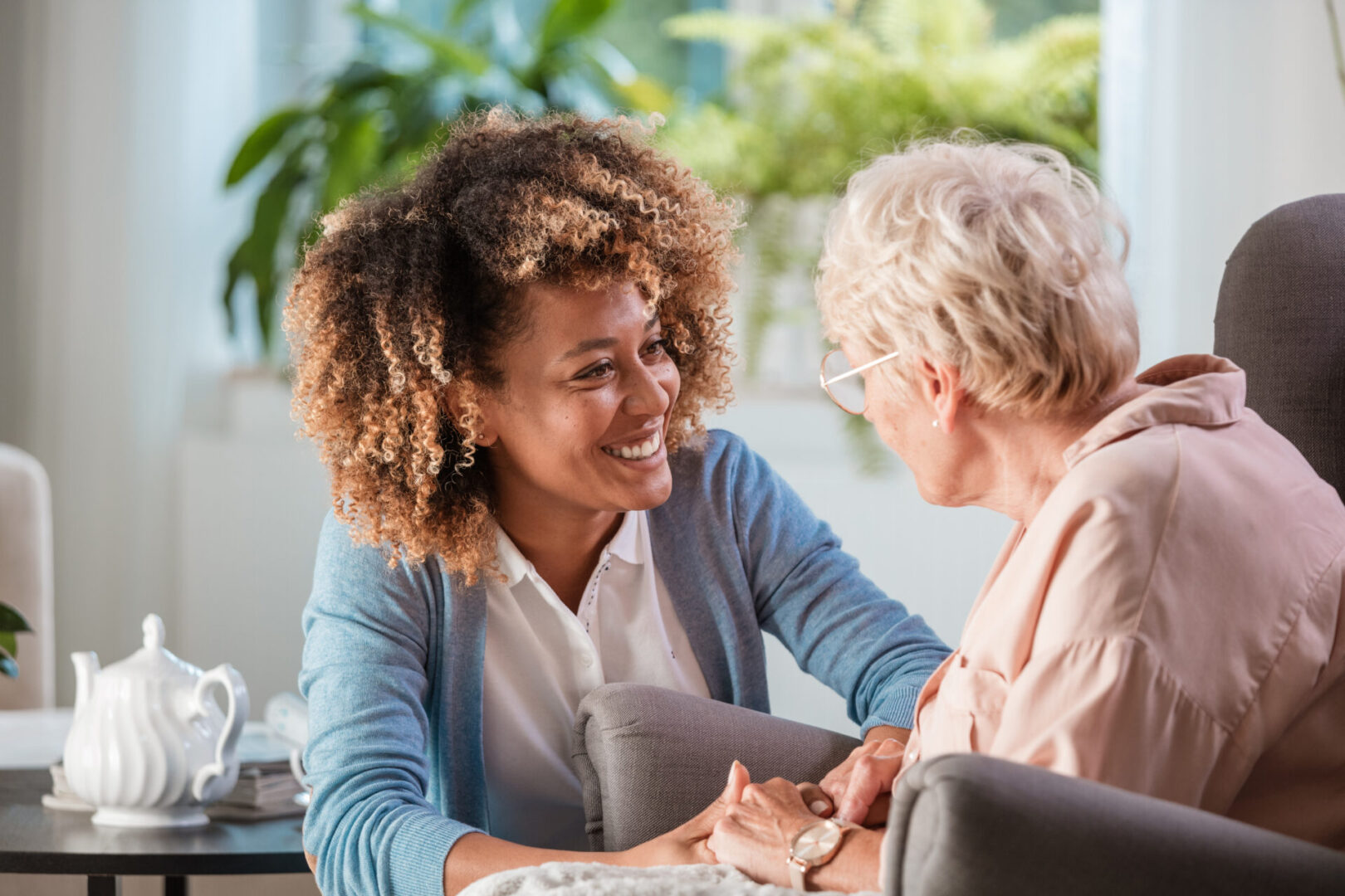 Smiling young female caregiver holding hands and talking with senior woman in living room